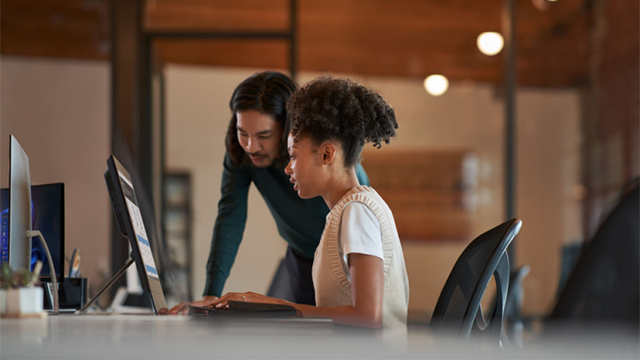 Two people in consultation while looking at a laptop. One person sits at desk; the other person is standing and leans over to look at the laptop.