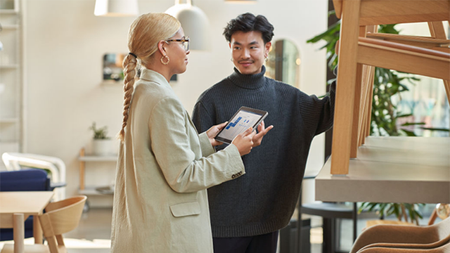 Two people--one holding a tablet--stand in a retail furniture showroom in conversation.