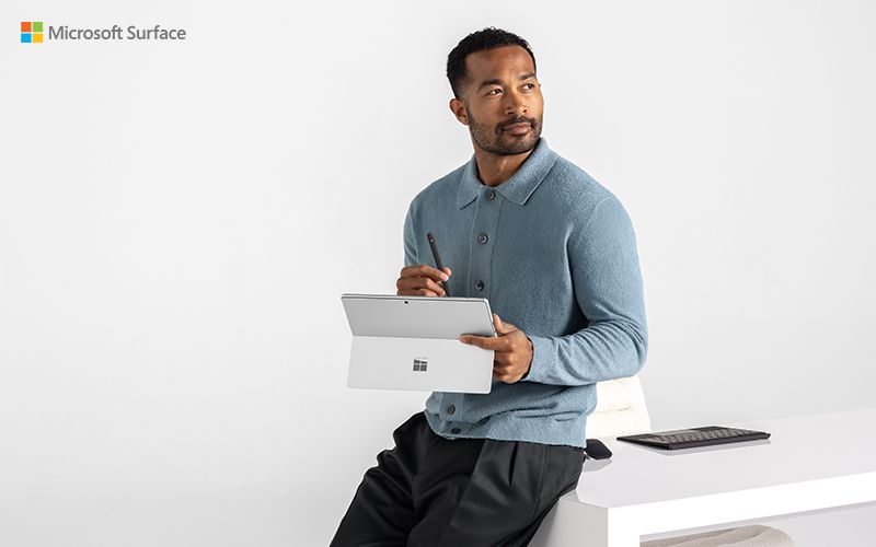 A person wearing a blue sweater leaning against a white desk. He's holding a Microsoft Surface Pro, and looking off to side, as if in deep thought.