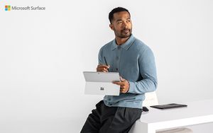 A person wearing a blue sweater leaning against a white desk. He's holding a Microsoft Surface Pro, and looking off to side, as if in deep thought.