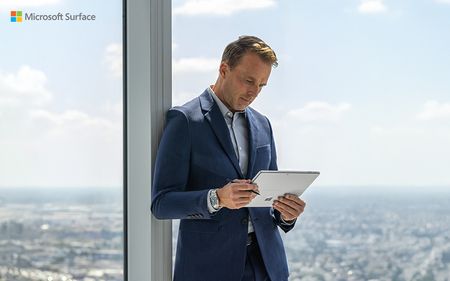 A person wearing a suit, holding a Microsoft Surface Pro, leaning against an office window.
