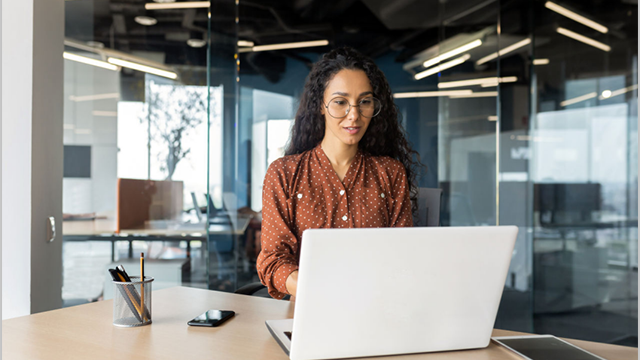 Woman in glass-walled office sits at a desk working on laptop.