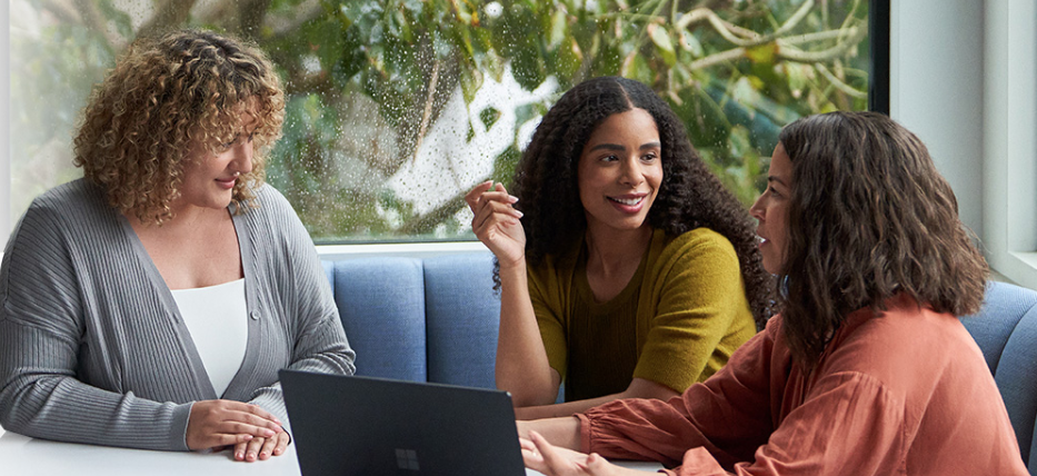Three individuals are seated around a table in a modern office setting, engaged in a discussion. One person is using a laptop, while the other two listen attentively. The background features large windows with raindrops on the glass and greenery outside.