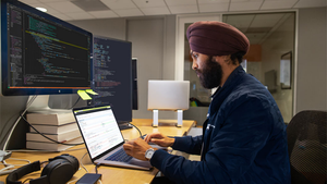 Person wearing a turban sits at a desk, working on a laptop with two additional monitors in front of him.