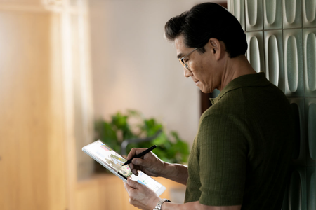 Person with short, dark hair in a green shirt and wearing glasses, using a Surface Pen on a Microsoft Surface Pro, leaning against a white wall with greenery in the background.