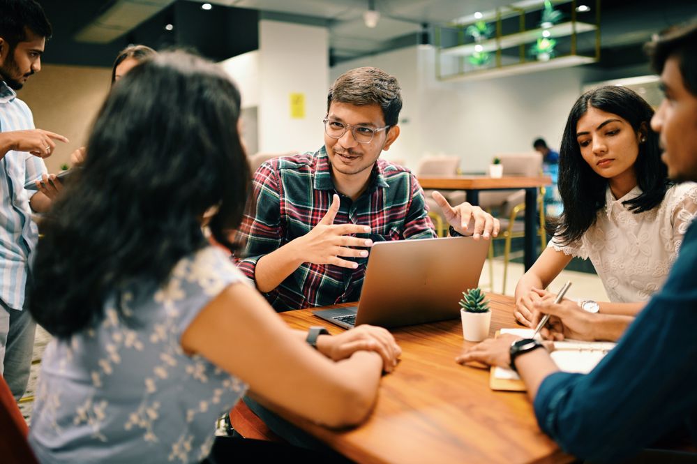 A group of individuals sitting at a table with one person in front of a laptop gesturing and talking to someone across from them.
