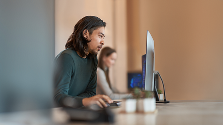 In the foreground of the photo, a man sits at a table working on a computer. In the background, a woman sits at the table working on a computer.