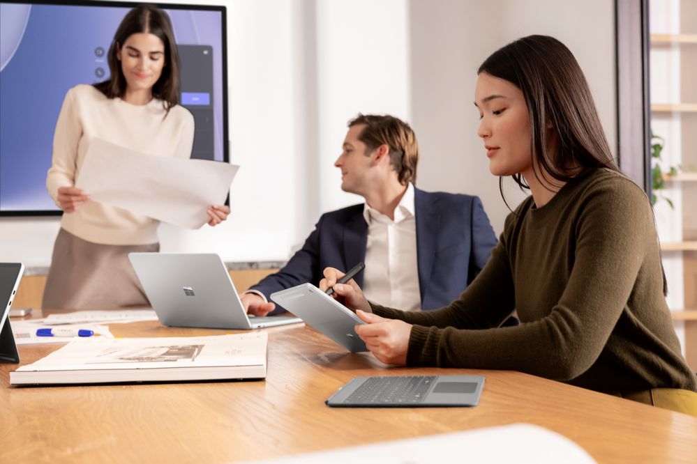 A woman holds a paper, a man is looking at the paper with her, and another woman is holding a Surface Pro 13 inch and a Surface Pen