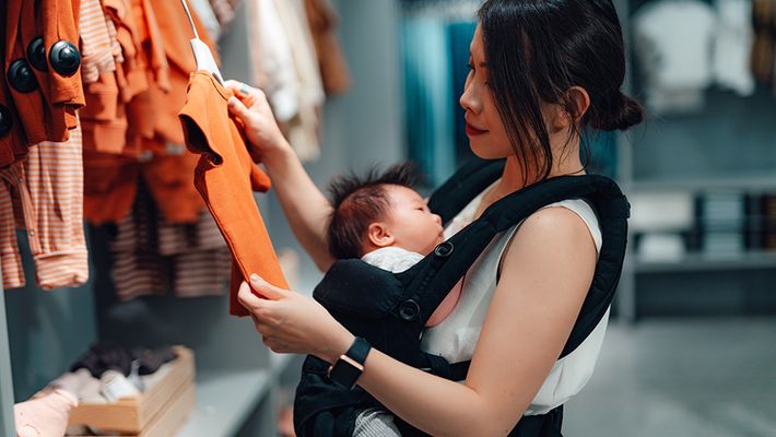 Mother choosing baby clothing while holding her baby in a baby carrier in a clothing store
