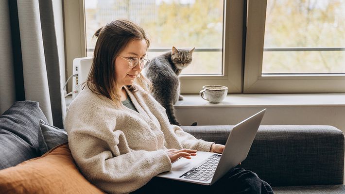 A woman works from home on a laptop while sitting on the sofa with her cat