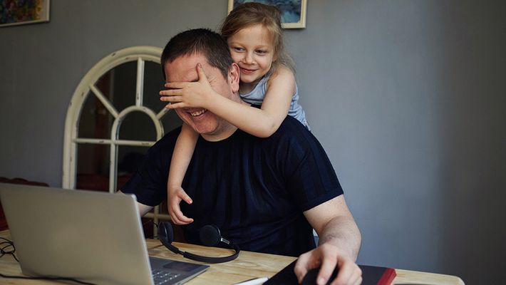 A little girl gives her father a hug while he's working on a laptop from home, playfully covering his eyes to stop him from working.
