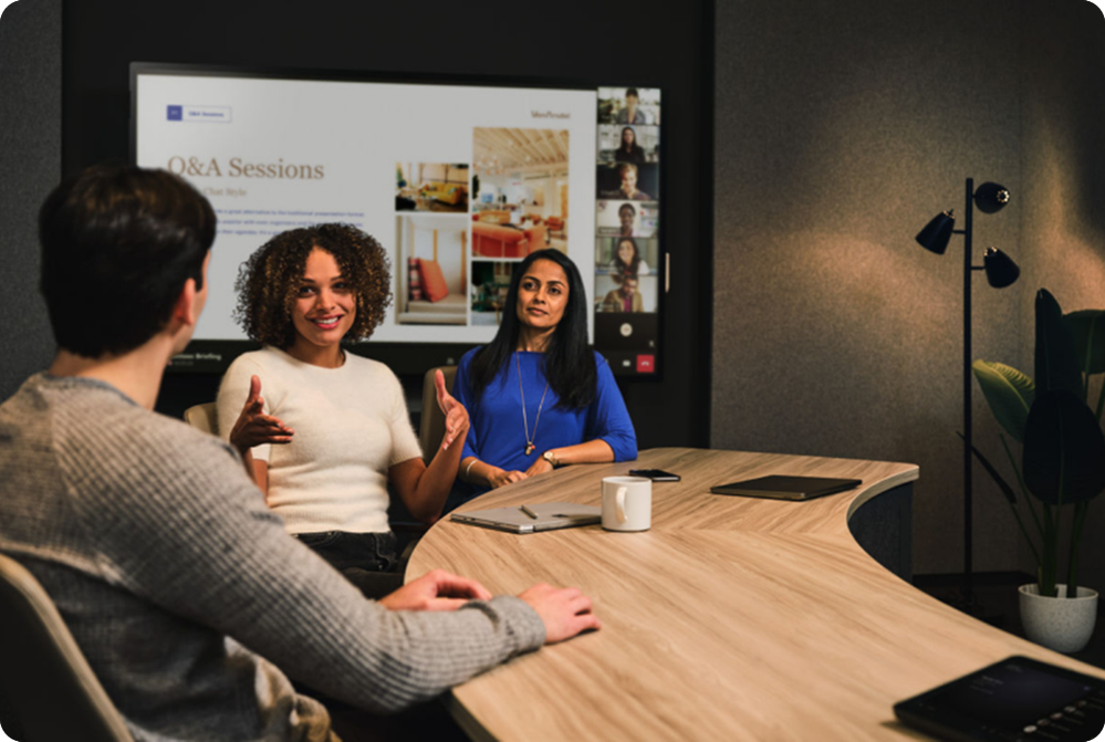 People in a meeting room sitting at a table that has a Teams Rooms on Windows touch board and console. The large display has a meeting slide in the background.