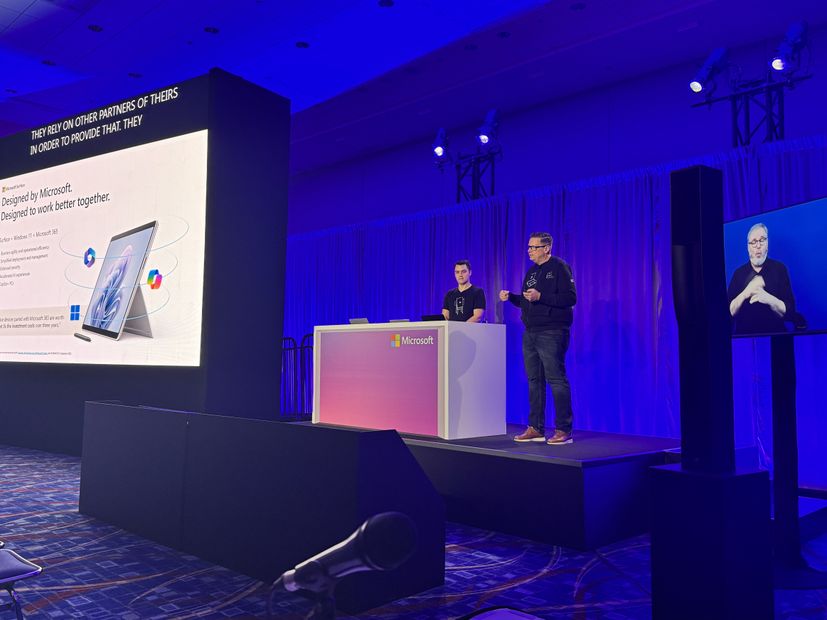 Two men on stage presenting with a picture of Surface on the screen to the left of stage