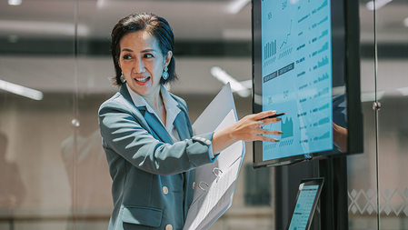 Businesswoman gestures at a screen displaying charts and graphs while presenting to an off-camera audience