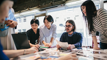 A group of people standing and sitting cowork at a conference table covered with papers pens and a laptop
