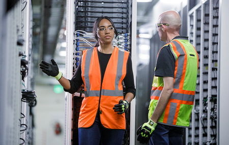 Two datacenter technicians wearing orange safety vests talking a datacenter, the woman gesturing at a rack of servers while the man listens.