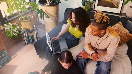 Three people sit on a sofa working on laptops in a sunlit room decorated with green plants, throw pillows, and artwork.