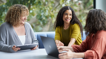 Three businesswomen meet at a corner conference table while working on a tablet and a laptop