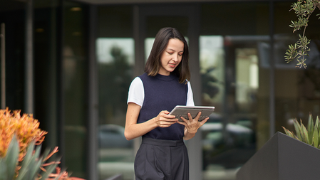 A woman working on a surface tablet walks through an office courtyard between two planters containing orange and green succulents