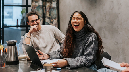 A woman wearing a gray textured sweater and a man wearing an off-white textured sweater collaborate at a conference table covered by a coffee urn, drink glasses, pens, papers, and a laptop. The man watches the woman, who is laughing.