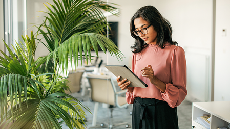 A businesswoman standing using a digital tablet in a modern office beside a leafy green majesty palm plant