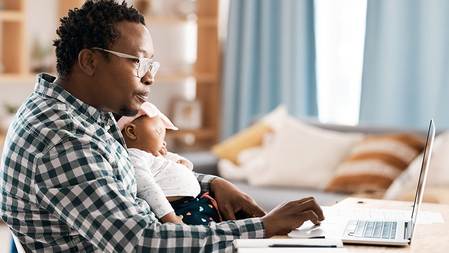 A man works on a laptop on a table while cradling a sleeping baby in his lap