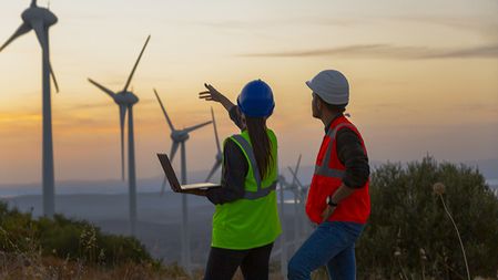 Two workers in helmets and safety vests work on a laptop and gesture at a wind turbine farm