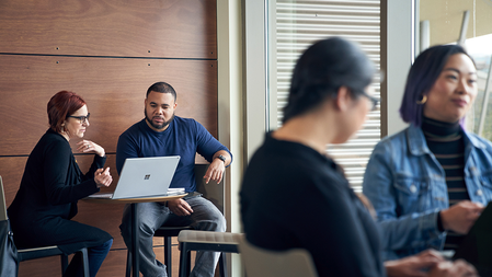 Man and a woman seated at a cafe table review data on a Surface laptop, while in the foreground two woman co-work at a cafe table