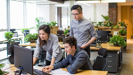 In an open office decorated with plants, a woman leans over a colleague's workstation to demonstrate something while he and another man observe