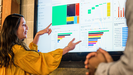 A woman presenting to colleagues gestures with both hands at a large display showing several multi-colored charts in different styles