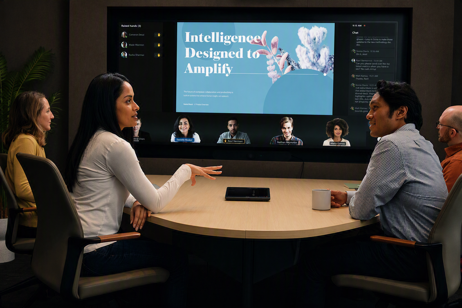 Four people sitting around a desk in a conference room, with a Teams Rooms system displaying a meeting on the display in the background
