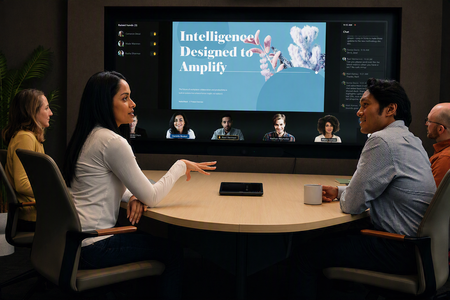 Four people sitting around a desk in a conference room, with a Teams Rooms system displaying a meeting on the display in the background