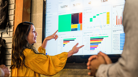 A woman shown in profile presents bar charts on a large display while a man, shown only by his hands, listens.