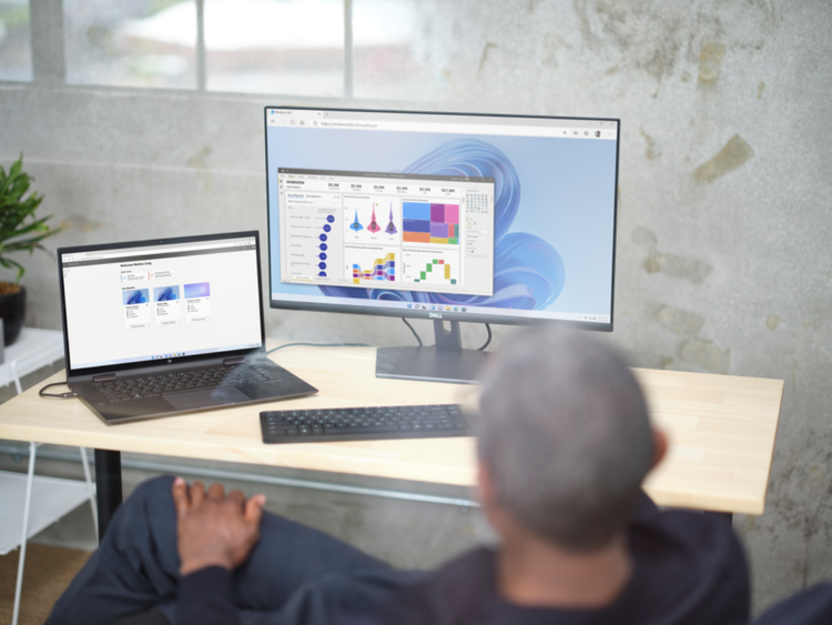 Photo of a man sitting at a desk with a laptop and monitor. The monitor shows a Windows 11 desktop while the laptop shows the Windows 365 Cloud PC selection screen